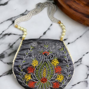 May include: A grey silk clutch bag with a floral embroidery design in shades of red, yellow, and green. The bag has a pearl and gold chain strap.