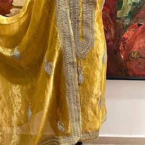 Puede incluir: Prenda de color amarillo mostaza con intrincados bordados plateados. La tela tiene una textura brillante y sedosa. El bordado se concentra a lo largo del escote y los bordes, con delicados motivos de cachemira. La prenda parece ser de estilo tradicional.