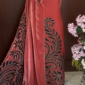 Puede incluir: Un dupatta de terciopelo rosa con diseños florales bordados en negro y un borde festoneado. El dupatta está drapeado sobre un vestido rosa con bordados similares.
