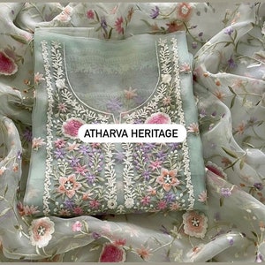 May include: A light green embroidered tunic with floral detailing in pink, purple, and white. The tunic is displayed on a matching floral patterned fabric. The text "ATHARVA HERITAGE" is visible on the tunic.