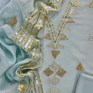 May include: A pale blue embroidered outfit with gold and silver detailing. The design includes geometric shapes and gold striped trim. The fabric looks lightweight and flowing, with a matching pale blue fabric square.