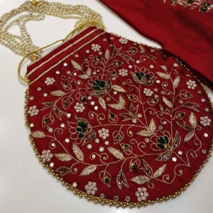 May include: A red velvet pouch with a round shape and a beaded trim. The pouch is decorated with gold and silver embroidery, including floral designs and sequins.