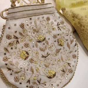 May include: A white fabric pouch with a gold beaded trim and intricate floral embroidery in gold and yellow thread. The pouch has a drawstring closure and is decorated with small, iridescent sequins.