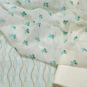May include: White fabric with a light blue floral pattern. The fabric is sheer and has a delicate texture. A second piece of white fabric with a wavy pattern is visible in the background.