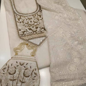 May include: An off-white embroidered outfit, comprising a tunic, trousers, and a scarf. The tunic showcases detailed floral and leaf designs in gold and white beads. The scarf is adorned with sequins, and the ensemble includes a matching purse.