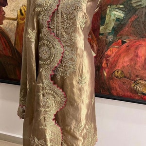 Puede incluir: Un vestido bordado de color dorado con ribete rosa. El vestido tiene un patrón floral y está hecho de una tela transparente.