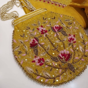 May include: A mustard yellow clutch bag with a beaded trim and floral embroidery. The embroidery features pink and red flowers with gold accents.