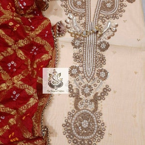 Puede incluir: Un vestido de color crema con intrincados bordados y abalorios. El vestido tiene un escote en V y una falda larga y fluida. La tela es fina y delicada. El vestido se combina con un dupatta rojo y dorado.