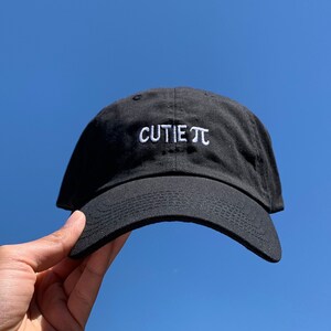 May include: A black baseball cap with white embroidery that reads "CUTIE π".