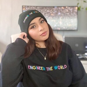 Pode incluir: Uma pessoa vestindo uma camisa de moletom preta com o texto "Engineer the World" em cores do arco-íris. Ela também está usando um gorro preto com o texto "Engineer the World" em cores do arco-íris.