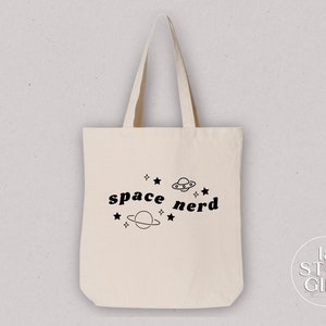 Op de afbeelding: Een witte canvas tote bag met zwarte tekst die "space nerd" zegt en een afbeelding van een planeet, sterren en een UFO.