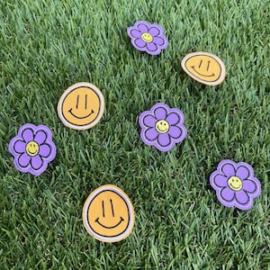 Puede incluir: Seis parches para planchar sobre una superficie de hierba verde. Hay tres parches amarillos con una cara sonriente y tres parches de flores moradas con caras sonrientes amarillas en el centro.