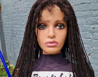 Può includere: Una testa di manichino con lunghe trecce marrone scuro. Il manichino indossa una maglietta viola scuro con la scritta "Beautyhub" e lo slogan "We make you look gorgeous and confident. We sell long lasting handmade wigs."