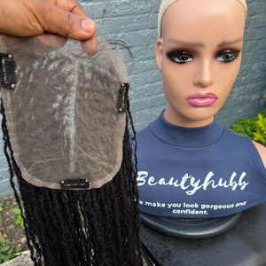 May include: A black wig with dreadlocks attached to a lace closure. The closure is made of a thin, transparent material and has clips on the sides. The wig is on a mannequin head with a blue shirt that says "Beautyhub" and "We make you look gorgeous and confident."