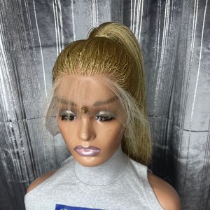May include: A blonde braided wig styled in a high ponytail, displayed on a mannequin head. The wig features a lace front and is worn with a gray top that has the text "Beautyhubb" and the words "we make you look gorgeous and confident".