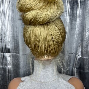 May include: A golden-blonde braided hair bun styled on a mannequin head. The bun is neatly arranged, with a textured appearance. The mannequin is wearing a gray turtleneck sweater. The background is a silver, reflective surface.