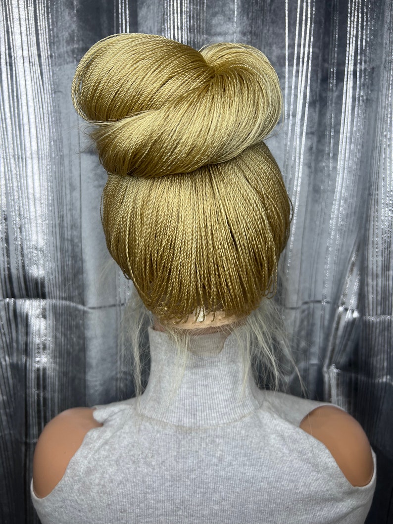 May include: A golden-blonde braided hair bun styled on a mannequin head. The bun is neatly arranged, with a textured appearance. The mannequin is wearing a gray turtleneck sweater. The background is a silver, reflective surface.