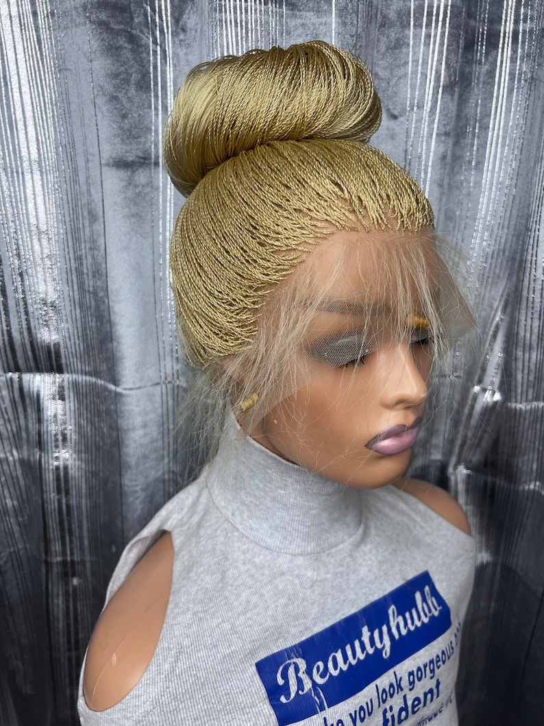 May include: A blonde braided wig styled in a high bun. The wig features a lace front and is worn over a gray turtleneck shirt with the text "Beautyhubb" on it. The background is a silver, textured surface.