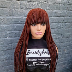 May include: A mannequin head displaying a long, braided wig in a warm auburn color. The wig features a blunt fringe and long, flowing braids. The mannequin is wearing a black shirt with the text "Beautyhub" and a slogan.