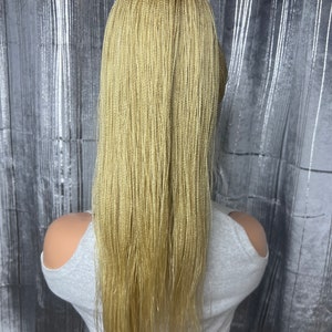 May include: A blonde micro braid wig with long, straight strands. The wig is styled on a mannequin head, showcasing the intricate braiding pattern and color. The background is a silver, textured curtain.