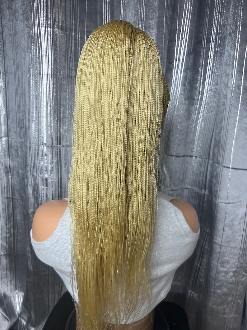 May include: A blonde micro braid wig with long, straight strands. The wig is styled on a mannequin head, showcasing the intricate braiding pattern and color. The background is a silver, textured curtain.