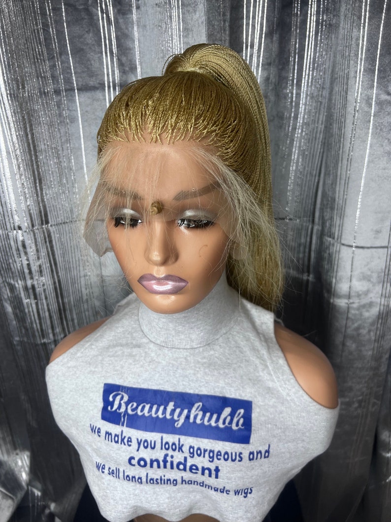 May include: A blonde braided wig styled in a high ponytail, displayed on a mannequin head. The wig features a lace front and is worn with a gray top that has the text "Beautyhubb" and the words "we make you look gorgeous and confident".