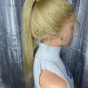 May include: A blonde micro-braided wig styled in a high ponytail. The wig features a lace front for a natural hairline and is secured on a mannequin head. The braids are a light, golden color, and the wig is worn with a gray turtleneck.