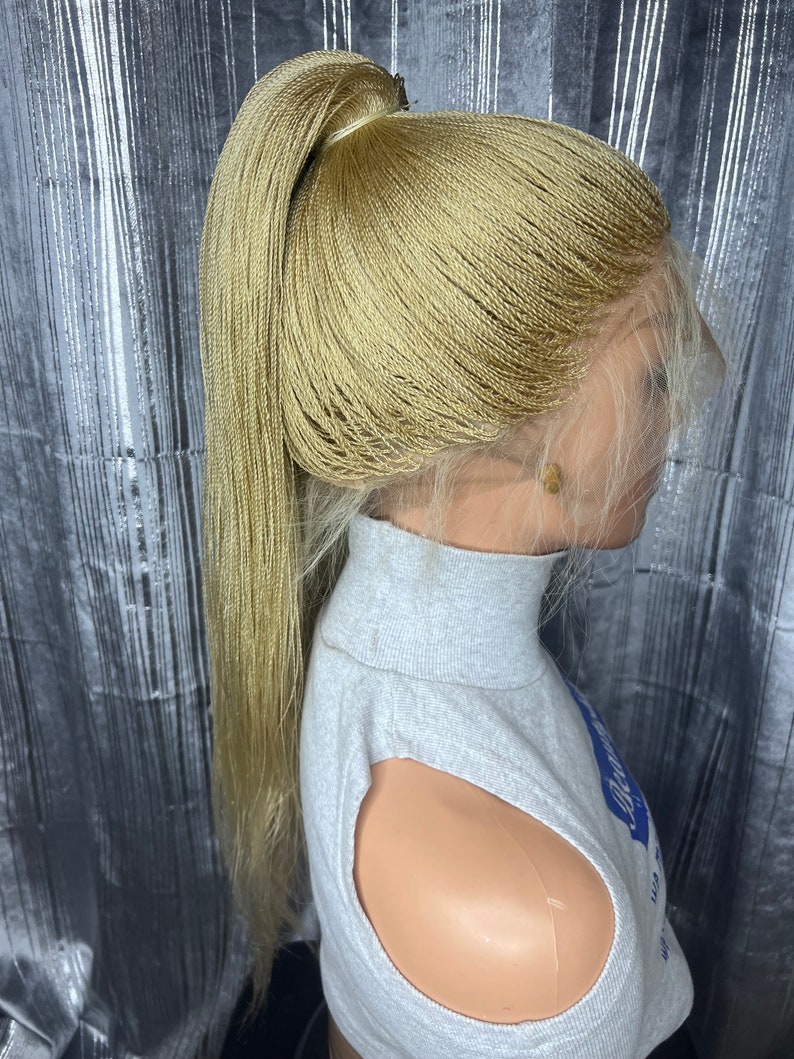 May include: A blonde micro-braided wig styled in a high ponytail. The wig features a lace front for a natural hairline and is secured on a mannequin head. The braids are a light, golden color, and the wig is worn with a gray turtleneck.