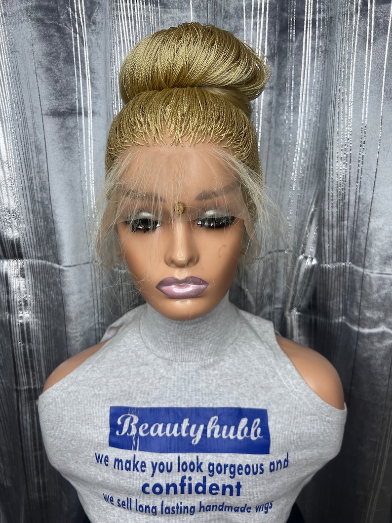 May include: A blonde wig styled in a high bun, displayed on a mannequin head. The wig features a lace front and a natural-looking hairline. The mannequin is wearing a gray top with the text "Beauty hubb" and the slogan "we make you look gorgeous and confident".