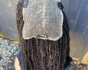 Può includere: Parrucca nera con dreadlocks, con chiusura in pizzo e clip per il fissaggio. La parrucca è esposta su una testa di manichino, evidenziando la consistenza e la lunghezza dei capelli. La chiusura in pizzo è beige chiaro.