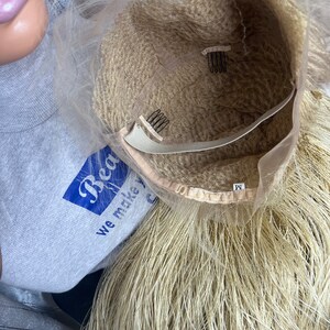 May include: A blonde wig with a mesh cap and attached combs for secure wear. The wig has long, straight strands and a light brown interior. The label on the wig reads "M".
