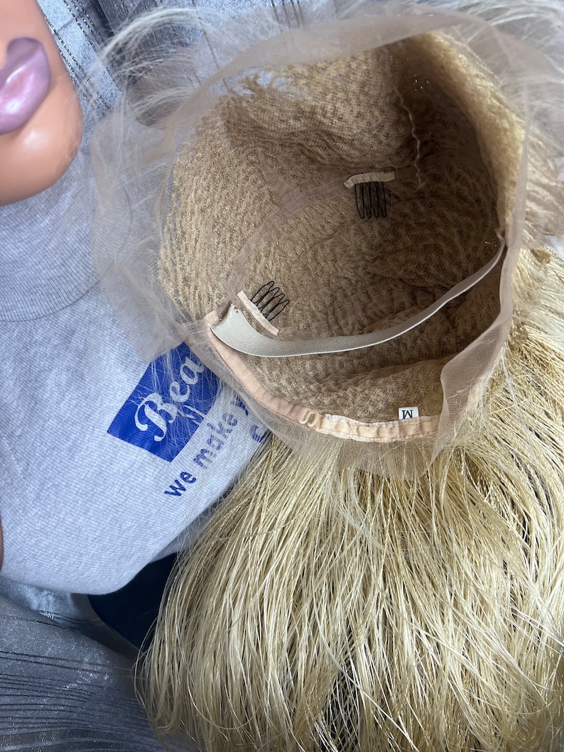 May include: A blonde wig with a mesh cap and attached combs for secure wear. The wig has long, straight strands and a light brown interior. The label on the wig reads "M".