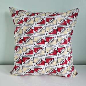 May include: Square throw pillow with a repeating pattern of red and cream-colored sneakers on a white background. The sneakers are outlined in black. The pillow is a decorative item for a bed or couch.