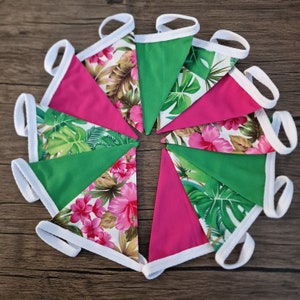 Op de afbeelding: Een guirlande van afwisselend roze en groene stoffen vlaggen met tropische blad- en bloemenpatronen. De vlaggen zijn aan elkaar genaaid met witte stof.