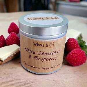 Może przedstawiać: Srebrny pojemnik z brązową etykietą z napisem "Wilson & Co. White Chocolate & Raspberry Hand crafted in Shropshire, England". Pojemnik jest otoczony świeżymi malinami.