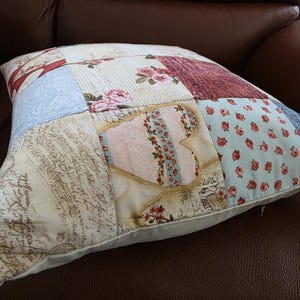 May include: A square throw pillow featuring a patchwork design with various fabric patterns including light blue with small red roses, cream with cursive script, pink hearts, and floral motifs in shades of red, pink, and blue. A zipper is visible on the side.