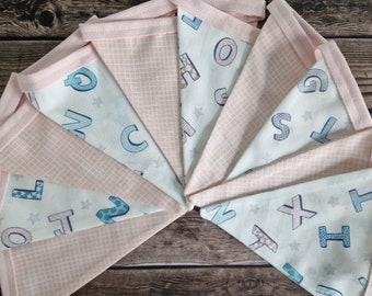 May include: A pink and white fabric banner with a light blue alphabet pattern. The banner is made of triangles with pink trim and features letters and stars.