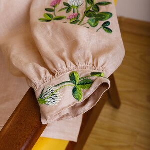 May include: A pale pink linen blouse with green and pink embroidered clover flowers. The blouse has a gathered cuff and is folded over a wooden armrest.