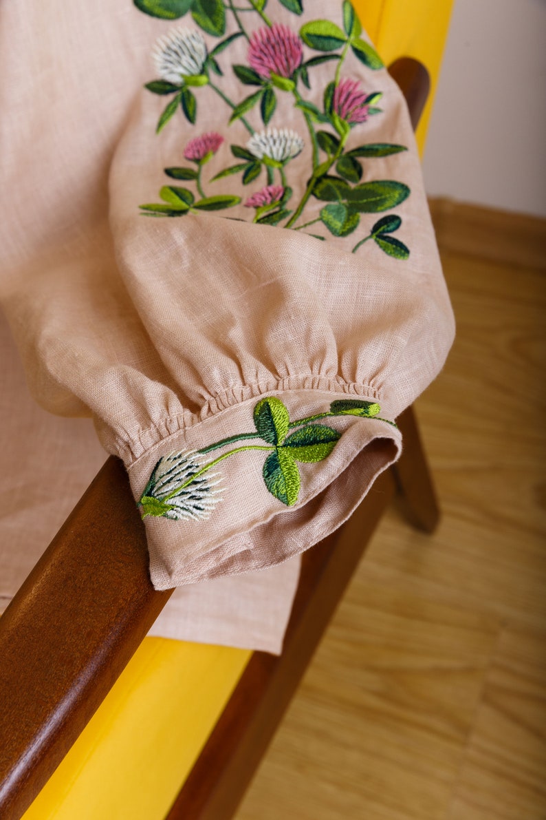 May include: A pale pink linen blouse with green and pink embroidered clover flowers. The blouse has a gathered cuff and is folded over a wooden armrest.
