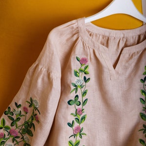 May include: A light beige linen blouse with embroidered clover flowers in shades of green, pink, and white. The blouse has a v-neck and is hanging on a white hanger.