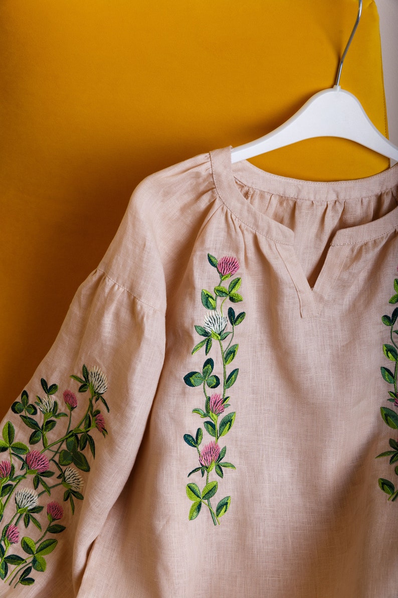 May include: A light beige linen blouse with embroidered clover flowers in shades of green, pink, and white. The blouse has a v-neck and is hanging on a white hanger.