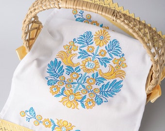 May include: A white linen kitchen towel with yellow and blue embroidered flowers and a yellow lace trim. The towel is folded and placed in a wicker basket.