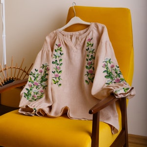 May include: A beige linen blouse with embroidered clover flowers in pink and green. The blouse has a round neckline and long sleeves. It is displayed on a yellow armchair.