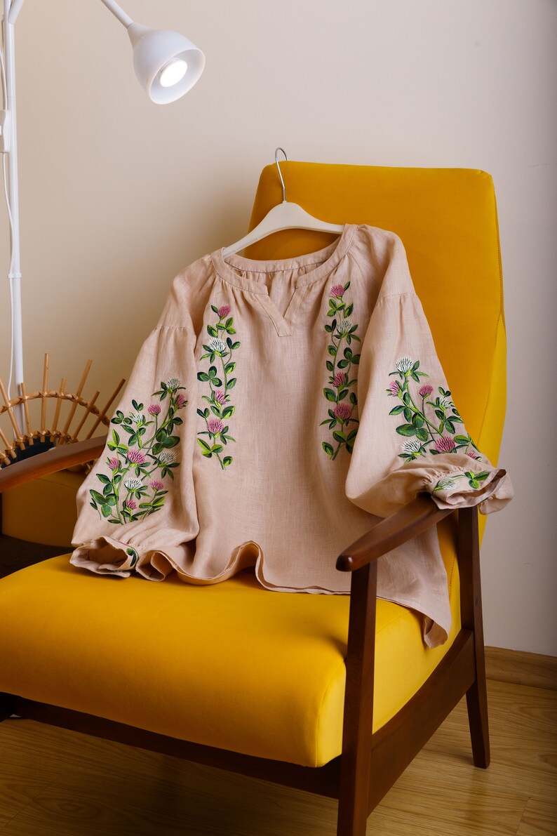 May include: A beige linen blouse with embroidered clover flowers in pink and green. The blouse has a round neckline and long sleeves. It is displayed on a yellow armchair.