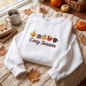 Op de afbeelding: Een wit sweatshirt met ronde hals en een geborduurd ontwerp van een geel esdoornblad, een stapel boeken, een oranje pompoen, een rood esdoornblad en een bruine koffiemok, met de bruine geborduurde tekst Cozy Season eronder geplaatst.