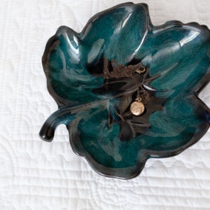 May include: A teal and black glazed ceramic leaf dish. A gold-toned necklace with an "Eva" pendant lies within the dish. The dish is set on a white, quilted surface. The dish is likely for storing jewellery.