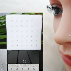 Op de afbeelding: Een close-up van een doorzichtige plastic container gevuld met kleine, zilveren neusringen met heldere edelstenen. De container is gelabeld met de nummers 1 tot en met 5. Een enkele neusring wordt getoond op een wit oppervlak met een liniaal voor schaal. Een persoon draagt een neusring met een heldere edelsteen.