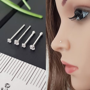 以下が含まれることがあります： 小さな透明な宝石がついた銀製の鼻のリングが4つあります。リングは黒い表面に並んでおり、背景には定規があります。画像の右側には、鼻のリングをつけた人がいます。
