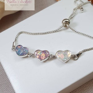 May include: A silver bracelet featuring three heart-shaped charms filled with colorful glitter. The charms are connected by a delicate chain, and the bracelet has an adjustable slider clasp. The brand name "Bella's lockets" is visible in the upper left corner.