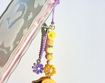 May include: A yellow and purple phone charm featuring a cartoon dog holding a blue heart. The charm is made of beads, flowers, and a small dog figurine.